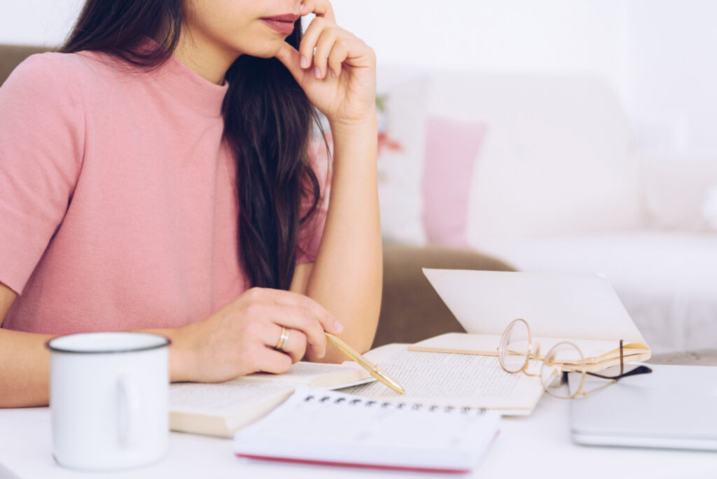 Woman sitting at a desk with notebooks, papers, coffee and glasses sitting on it.