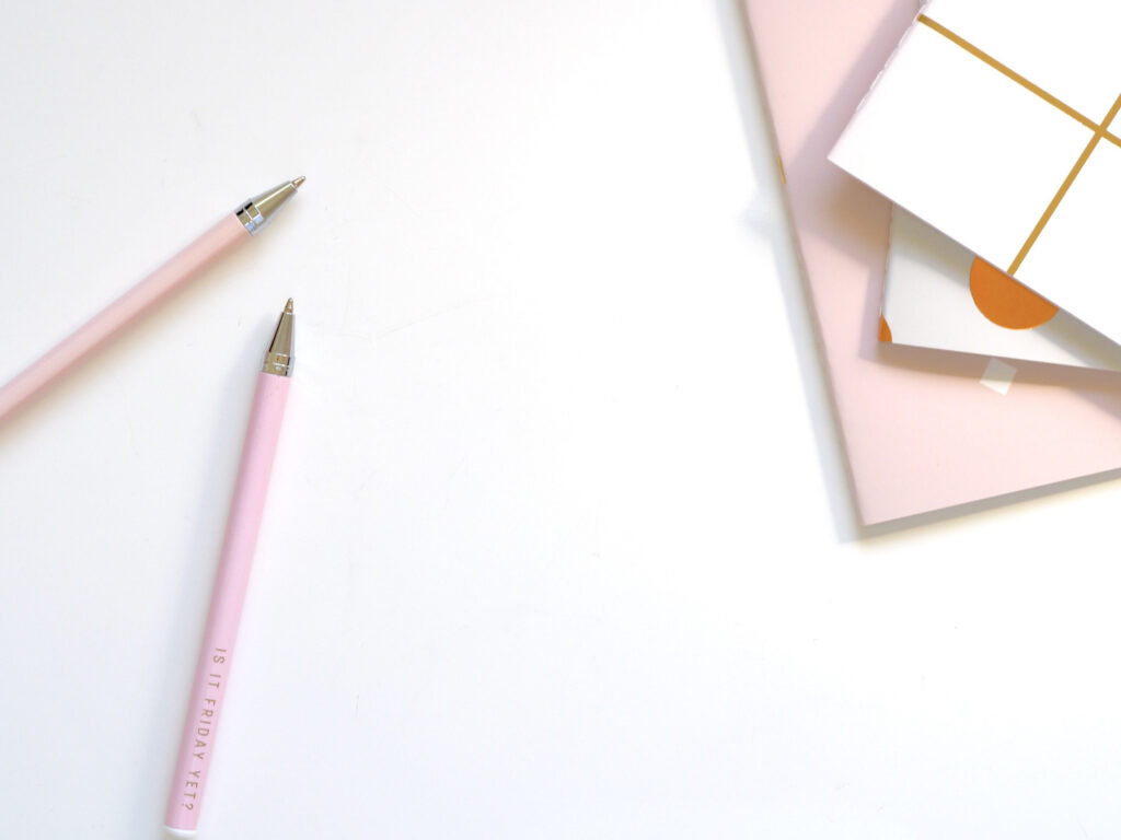 A white desk has pink pens and pink and white notebooks sitting on it.