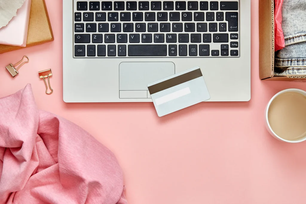 Top down view of a pink desk and a laptop with a credit card on top of it. There are other items scattered on the desk like a coffee mug, shipping box, and binder clips.