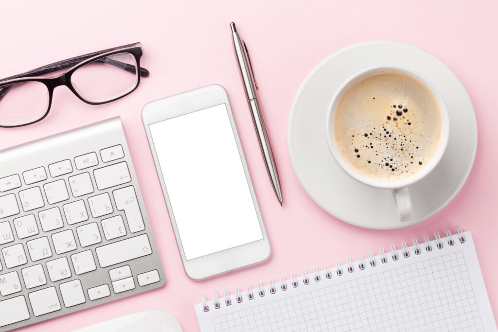 A pink desk has a laptop, cell phone, pen, latte and coffee cup on top of it.